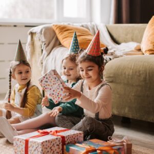 Three children happily opening wrapped birthday presents while wearing colorful party hats indoors.